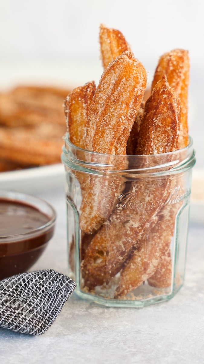 churros in glass jar with chocolate sauce and platter of churros in background.