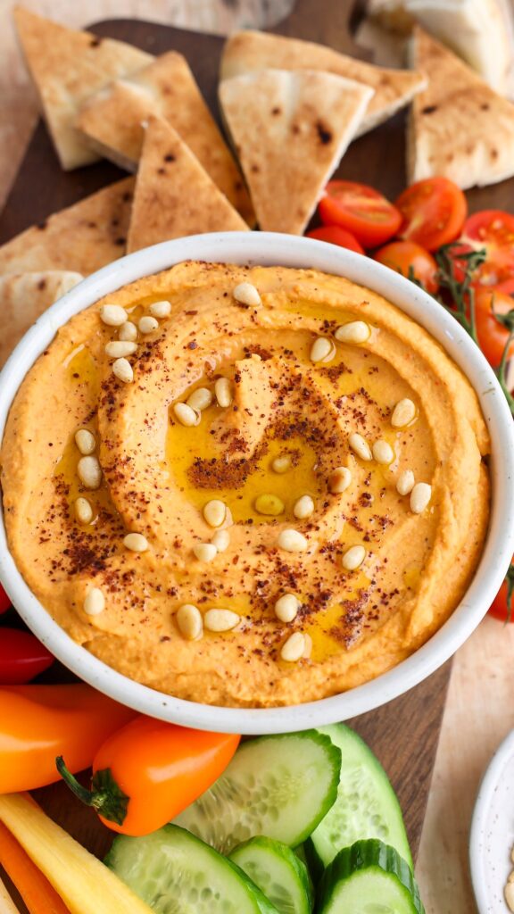 hummus topped with pine nuts, sumac, and olive oil in white serving bowl; bowl is placed between fresh vegetables and pita triangles