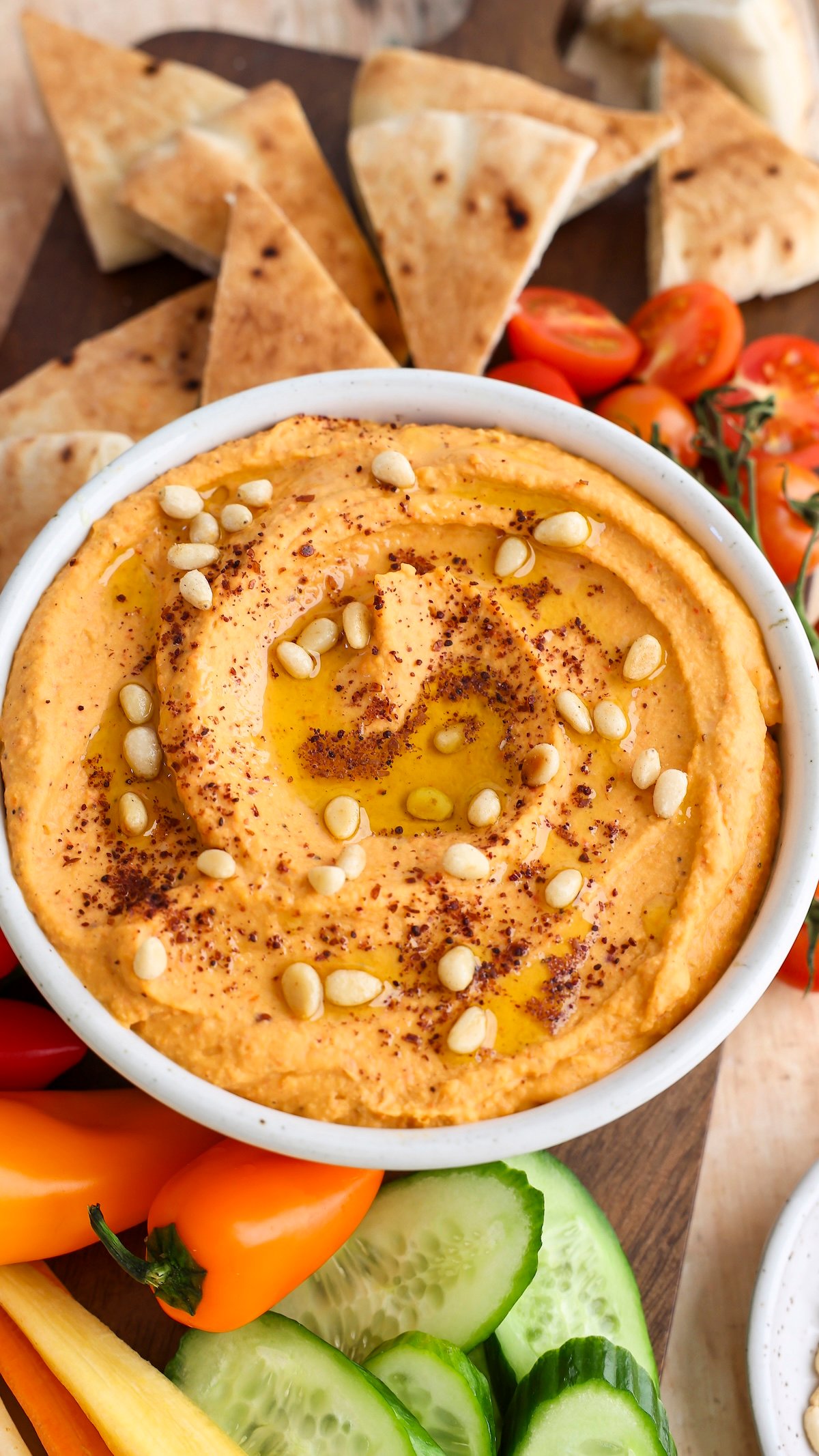 hummus topped with pine nuts, sumac, and olive oil in white serving bowl; bowl is placed between fresh vegetables and pita triangles