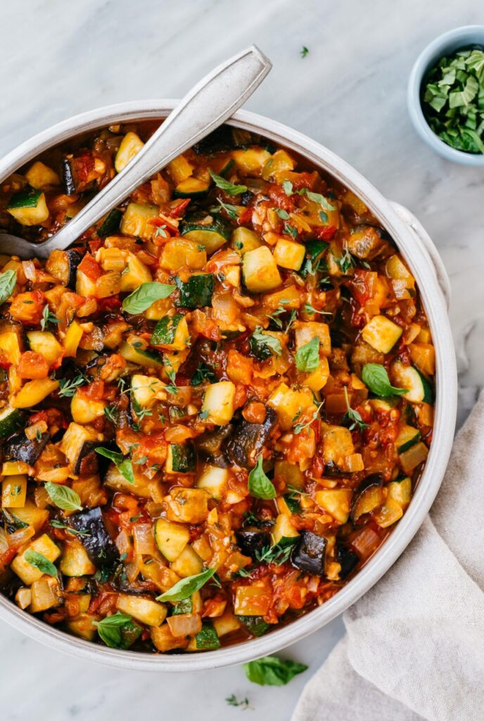 bowl of ratatouille with spoon and small bowl of fresh basil on the side.