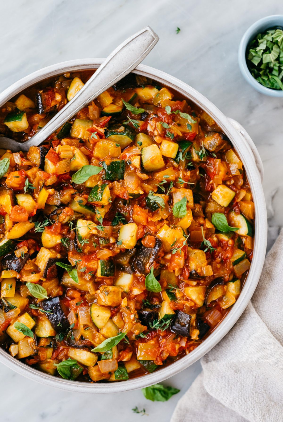 bowl of ratatouille with spoon and small bowl of fresh basil on the side.